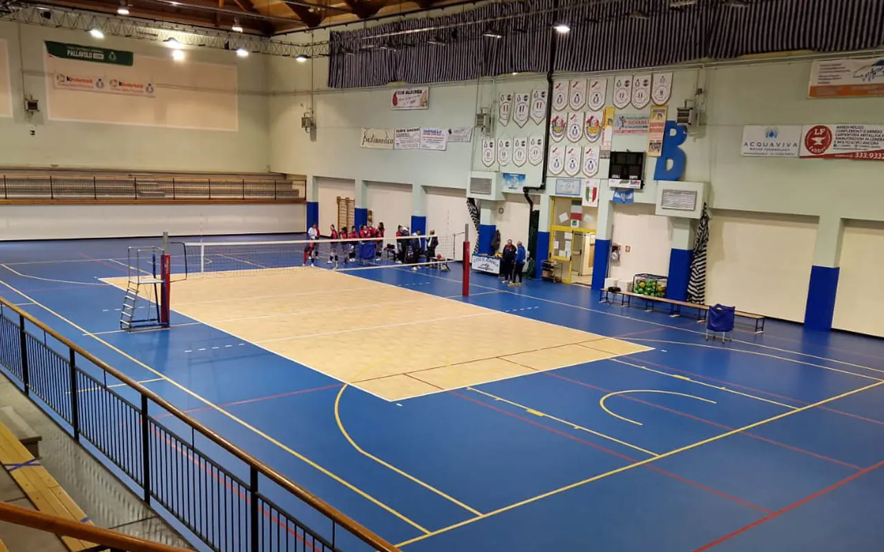 Mavi PVC zeminli kapalı salonda voleybol sahası, file direkleri ve ahşap tonlu oyun alanı