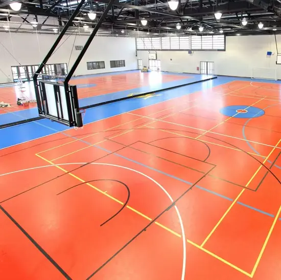 Kapalı spor salonunda kiremit ve mavi renkli PVC zemin, çok branşlı saha çizgileri ve basketbol potası